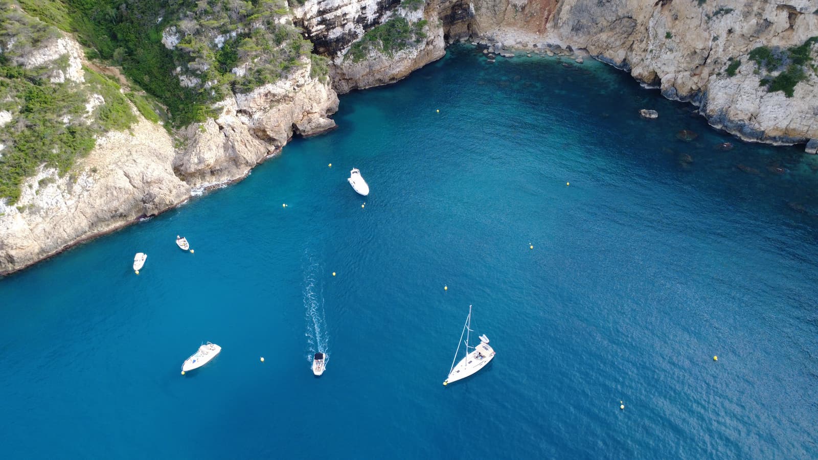 Vista aérea de una cala de la Costa Blanca con aguas turquesas, acantilados y varios barcos fondeados.